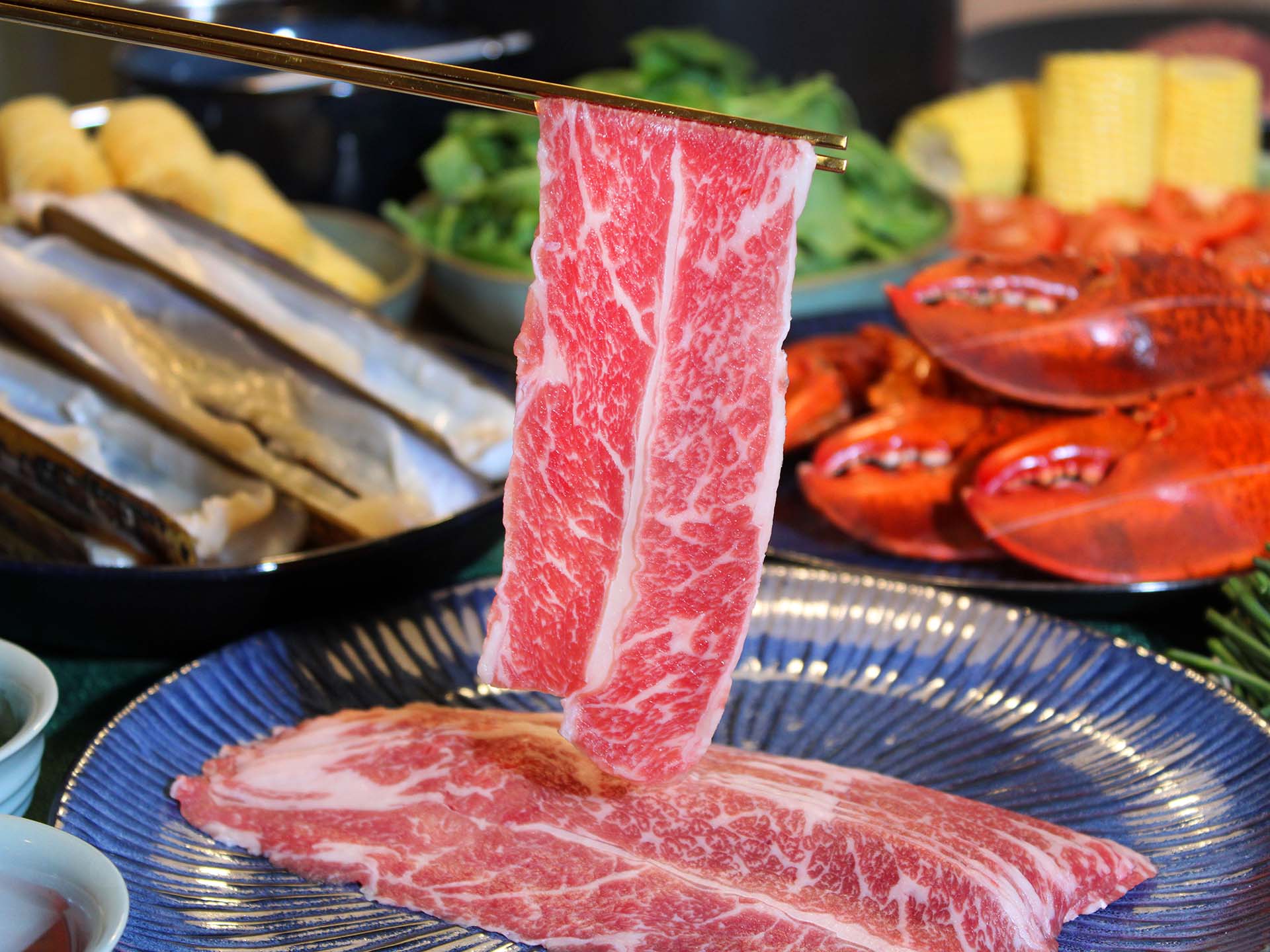 Raw beef slices on a blue plate with a background of cooked lobsters and vegetables.