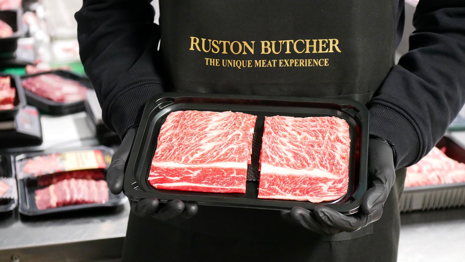 Person holding a package of boneless short ribs with 'Ruston Butcher' apron in a market setting