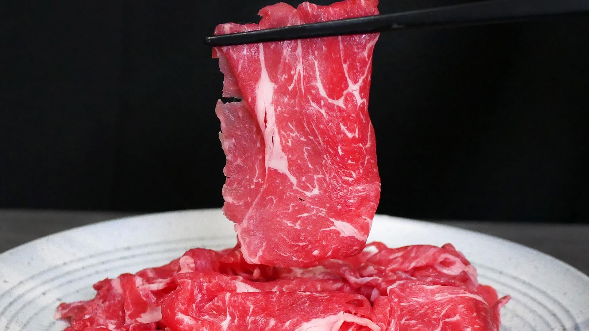 Raw beef slices being lifted with chopsticks on a white plate against a black background